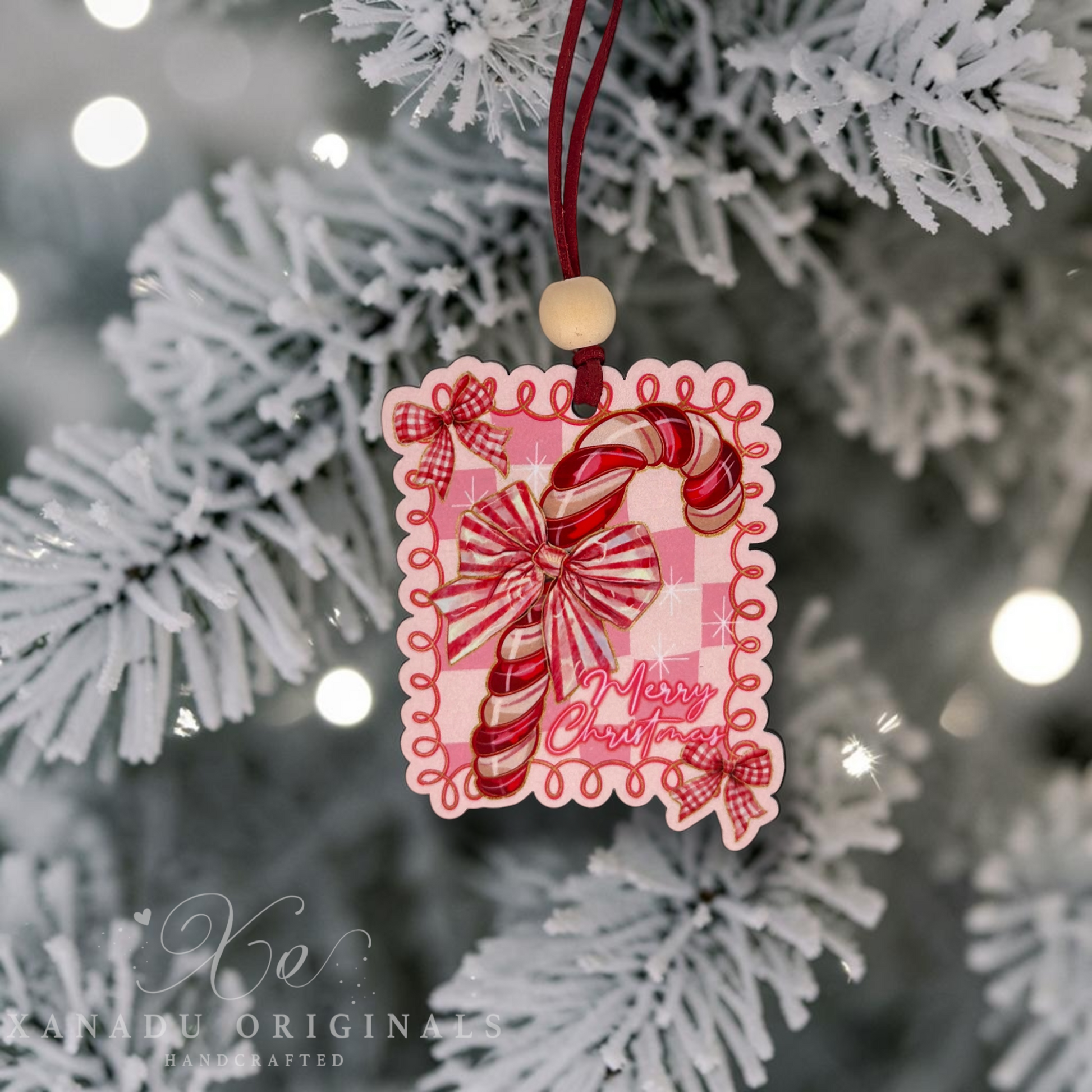 A square Christmas ornament with a scalloped pink border, featuring a candy cane, red bows, and the words 'Merry Christmas' on a pink checkered background, hanging from a red string on a white frosted Christmas tree.