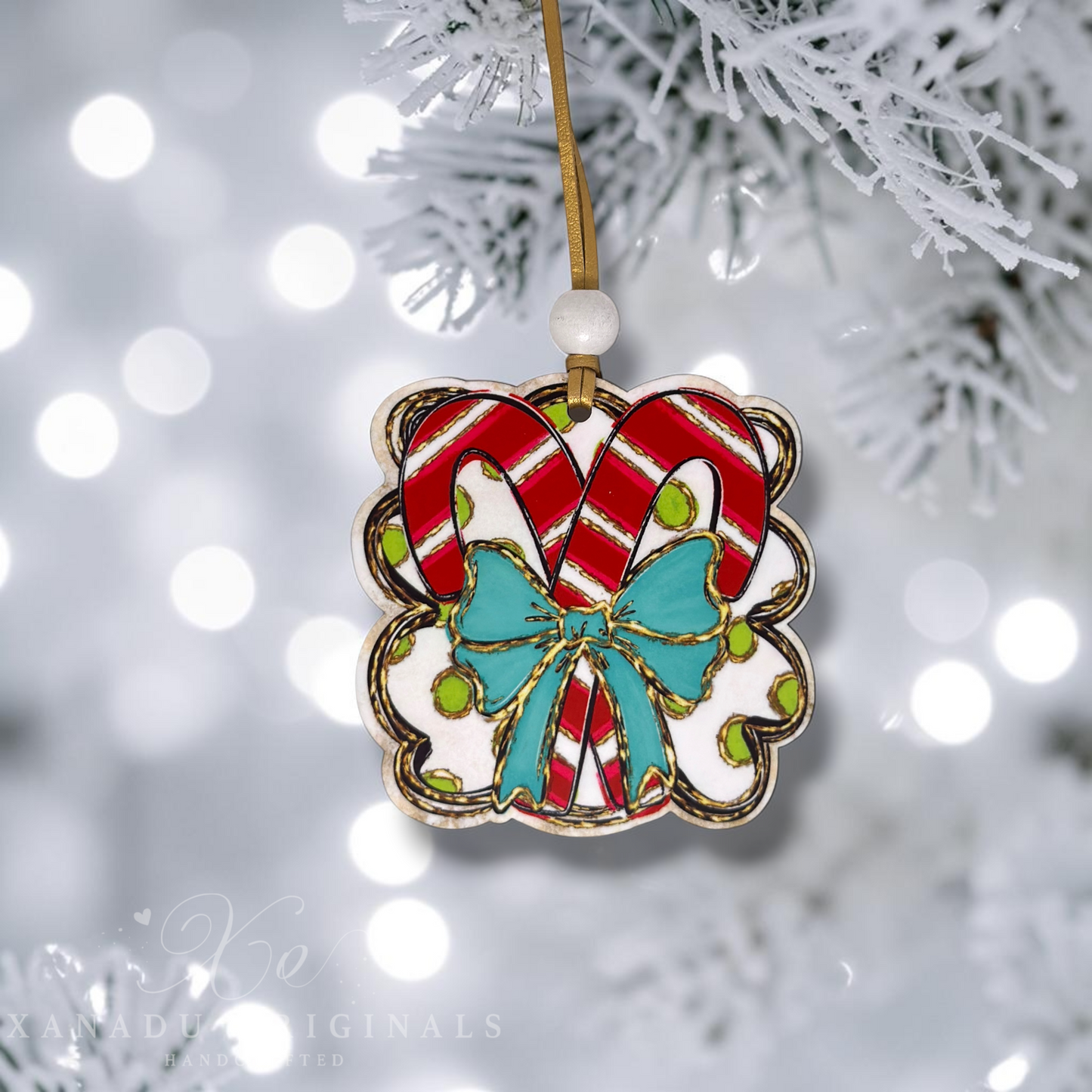 A scalloped ceramic Christmas ornament featuring two red-and-white striped candy canes and a turquoise bow, hanging from a gold string on a frosted tree branch with soft bokeh lights in the background.