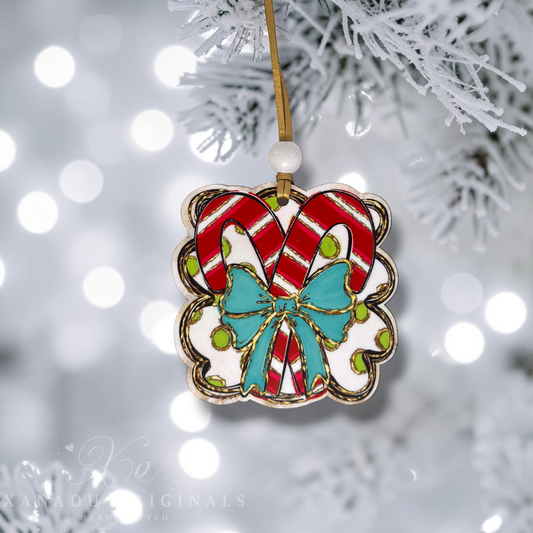 A scalloped ceramic Christmas ornament featuring two red-and-white striped candy canes and a turquoise bow, hanging from a gold string on a frosted tree branch with soft bokeh lights in the background.