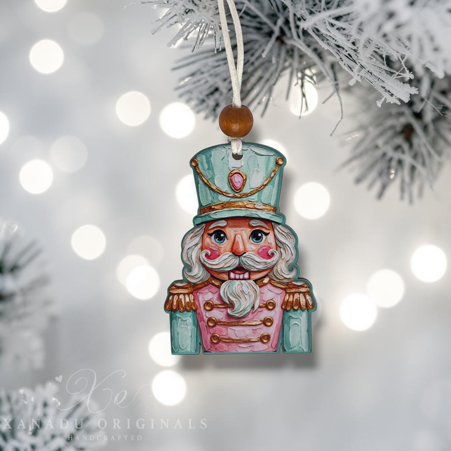 Handcrafted painted nutcracker Christmas ornament hanging from a white string on a frosted pine branch, with soft white bokeh lights in the background. The ornament is painted in light blue, pink, and white with gold accents.