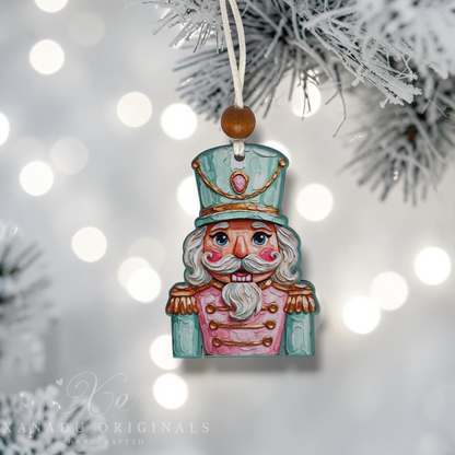 Handcrafted painted nutcracker Christmas ornament hanging from a white string on a frosted pine branch, with soft white bokeh lights in the background. The ornament is painted in light blue, pink, and white with gold accents.