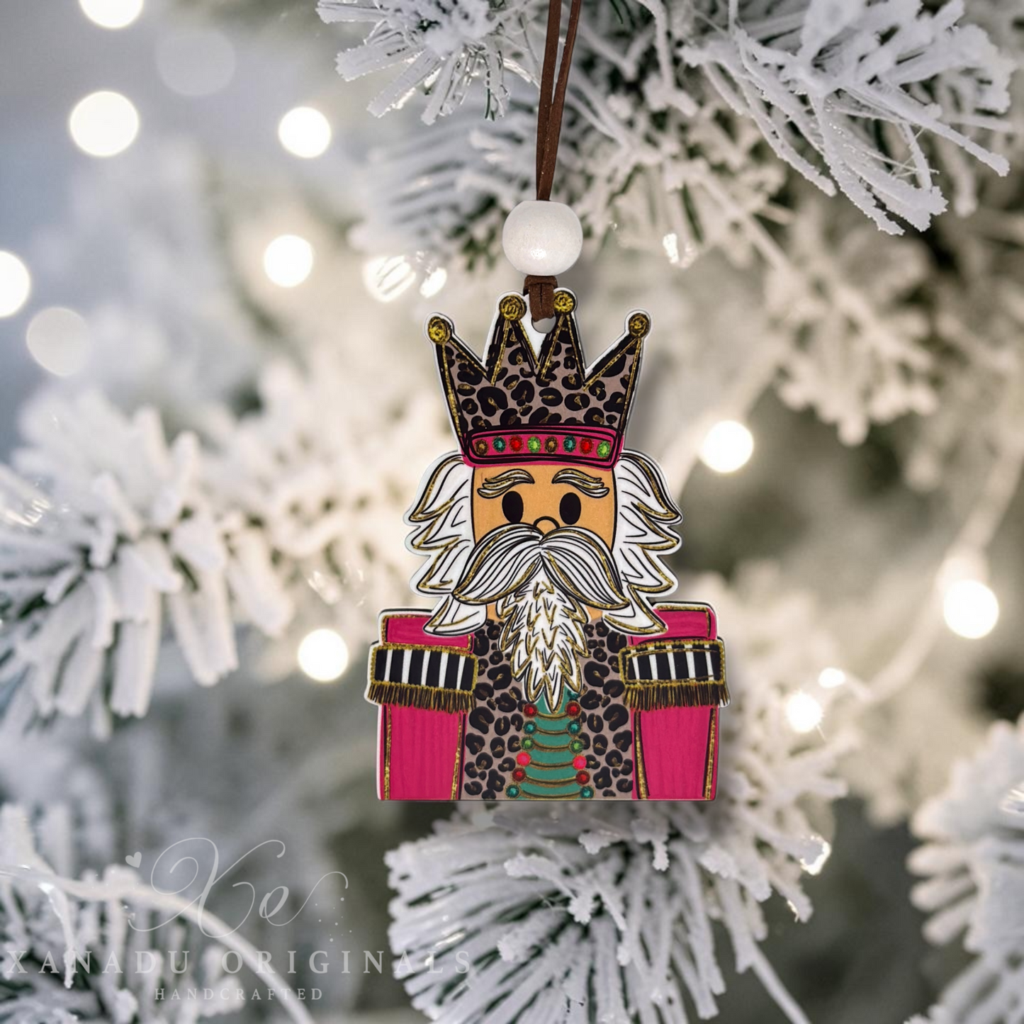 A handcrafted wooden Christmas nutcracker ornament featuring a leopard-print crown, white beard, and pink arms, hanging on a white-frosted Christmas tree with warm bokeh lights.