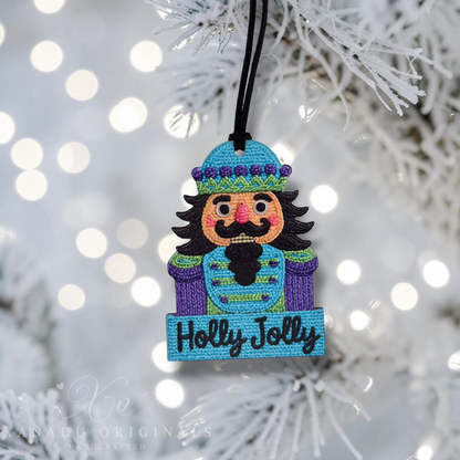 A handmade embroidered Christmas nutcracker ornament hanging from a white frosted Christmas tree branch. The ornament features a blue and purple nutcracker figure with a black beard and mustache, and a blue banner at the bottom embroidered with 'Holly Jolly' in black.