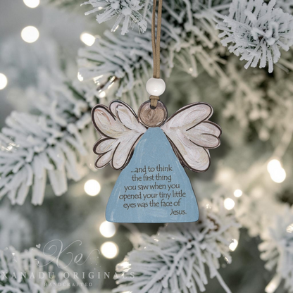A handcrafted wooden angel Christmas ornament with white wings and a light blue body, hanging from a string in front of a blurred flocked Christmas tree with warm string lights. The body of the ornament features a printed quote about Jesus.