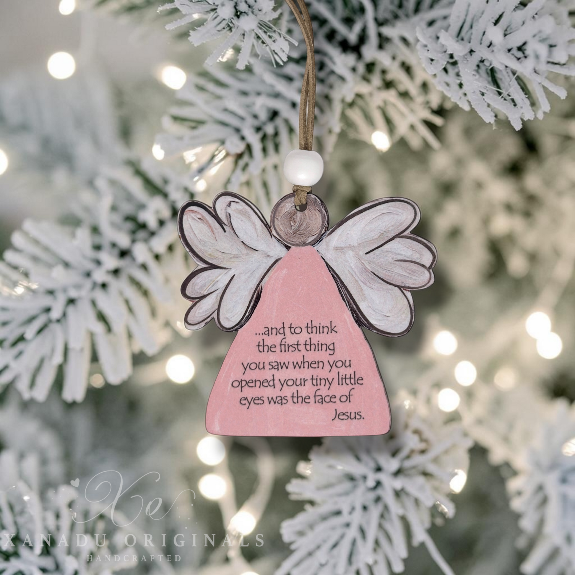 A pink angel-shaped Christmas ornament with white wings and a hanging loop, displayed on a blurred flocked Christmas tree with warm bokeh lights. The ornament has a printed message about a baby born sleeping and Jesus.