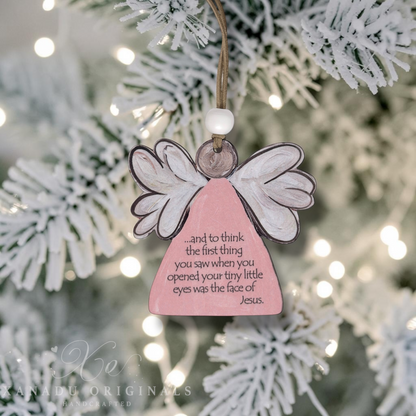 A pink angel-shaped Christmas ornament with white wings and a hanging loop, displayed on a blurred flocked Christmas tree with warm bokeh lights. The ornament has a printed message about a baby born sleeping and Jesus.
