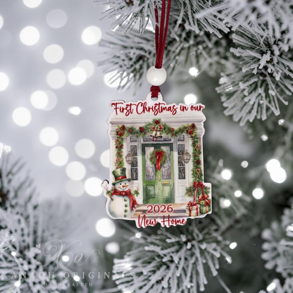 A personalized Christmas ornament shaped like a house door, hanging from a red ribbon on a snowy pine tree. The ornament features a green front door, red lettering that reads 'First Christmas in our 2026 New Home,' a snowman on the left, and wrapped gifts at the bottom.