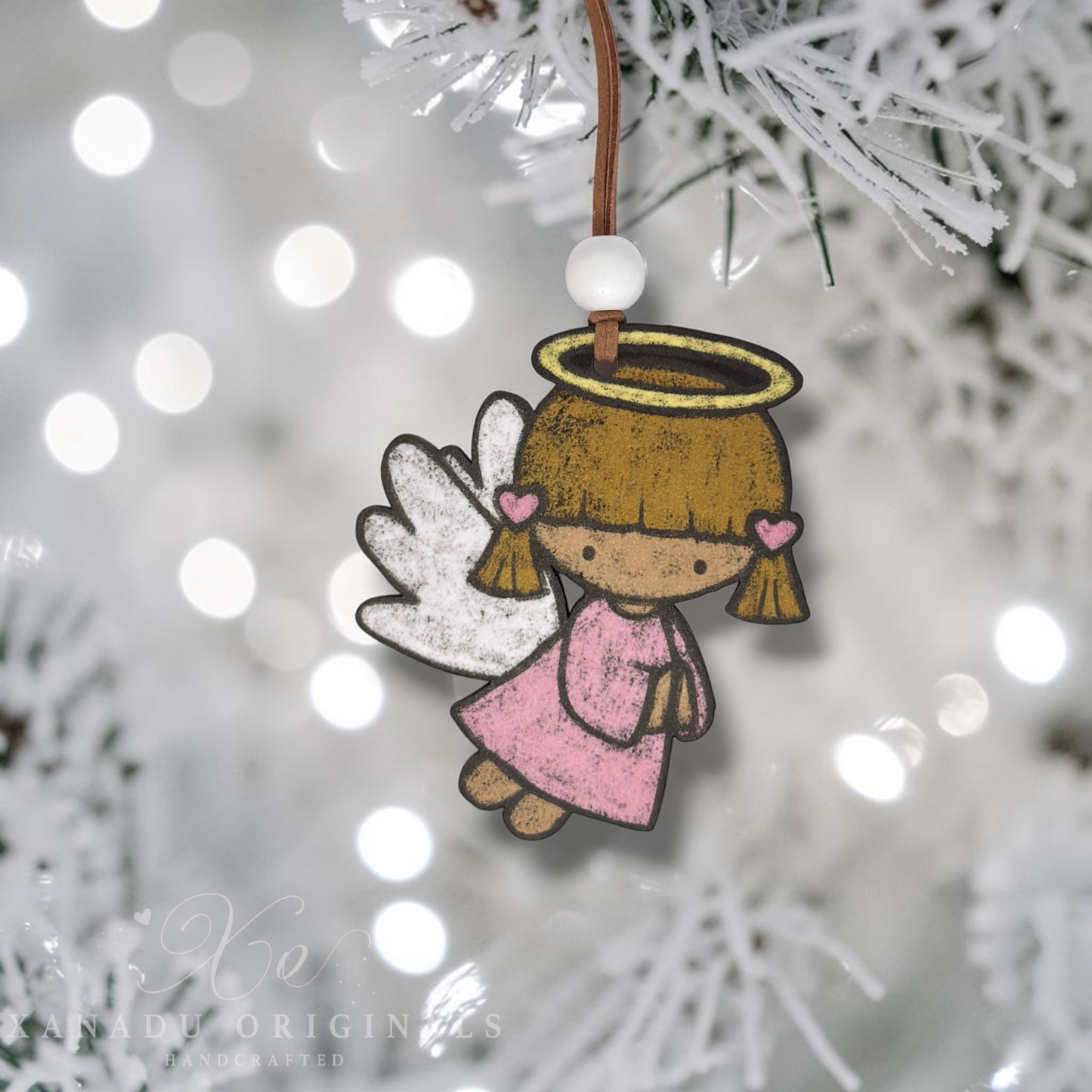 A handcrafted paper angel Christmas ornament with a brown hair halo, pink dress, and white wings, hanging from a brown string with a white bead against a blurred white Christmas tree background with bokeh lights.