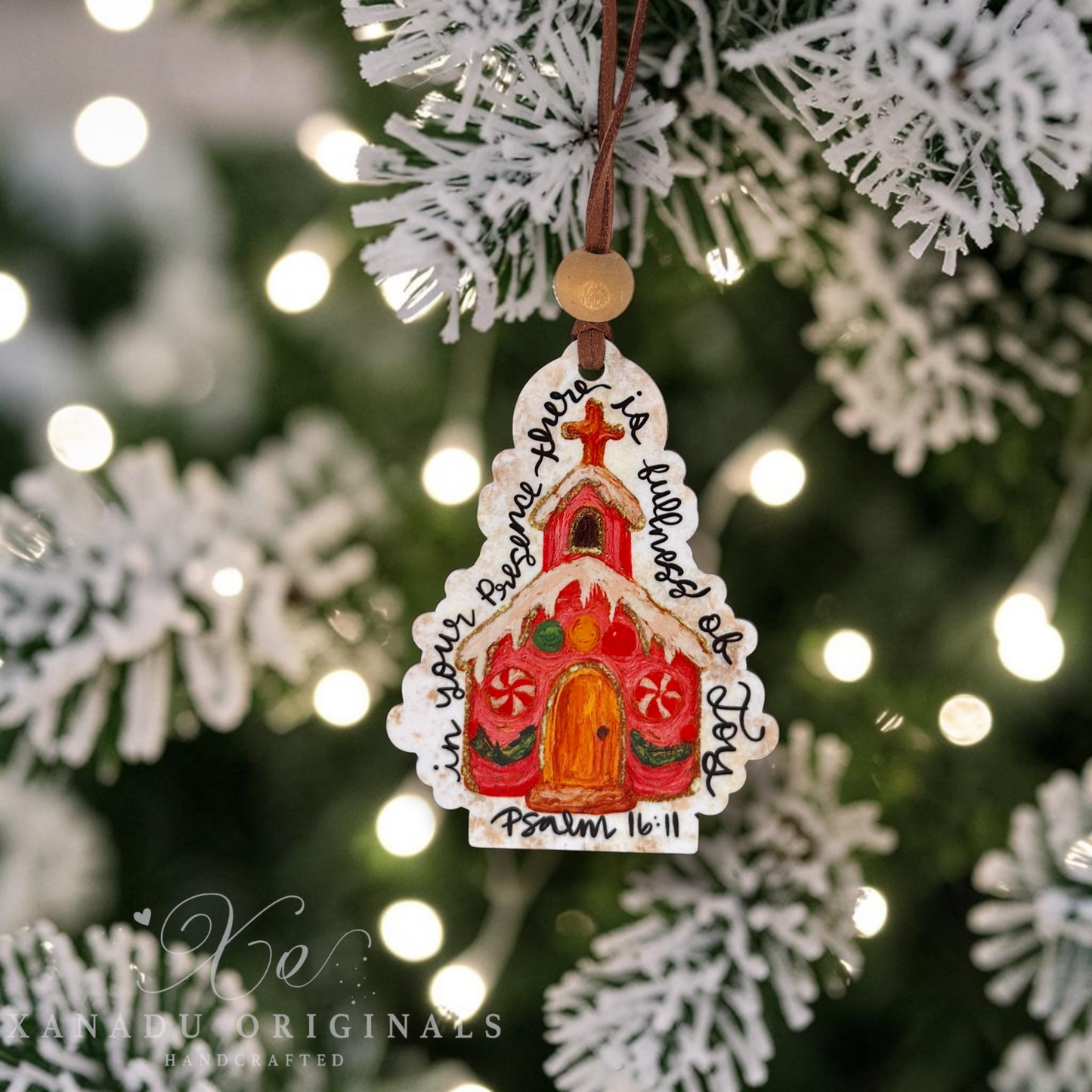 A hand-painted wooden Christmas ornament shaped like a church hangs from a brown string on a decorated Christmas tree with white artificial branches and warm bokeh lights. The ornament features a colorful painted church with a red roof, green accents, a yellow door, and black handwritten text reading 'in your presence there is fullness of joy' Psalm 16:11'.