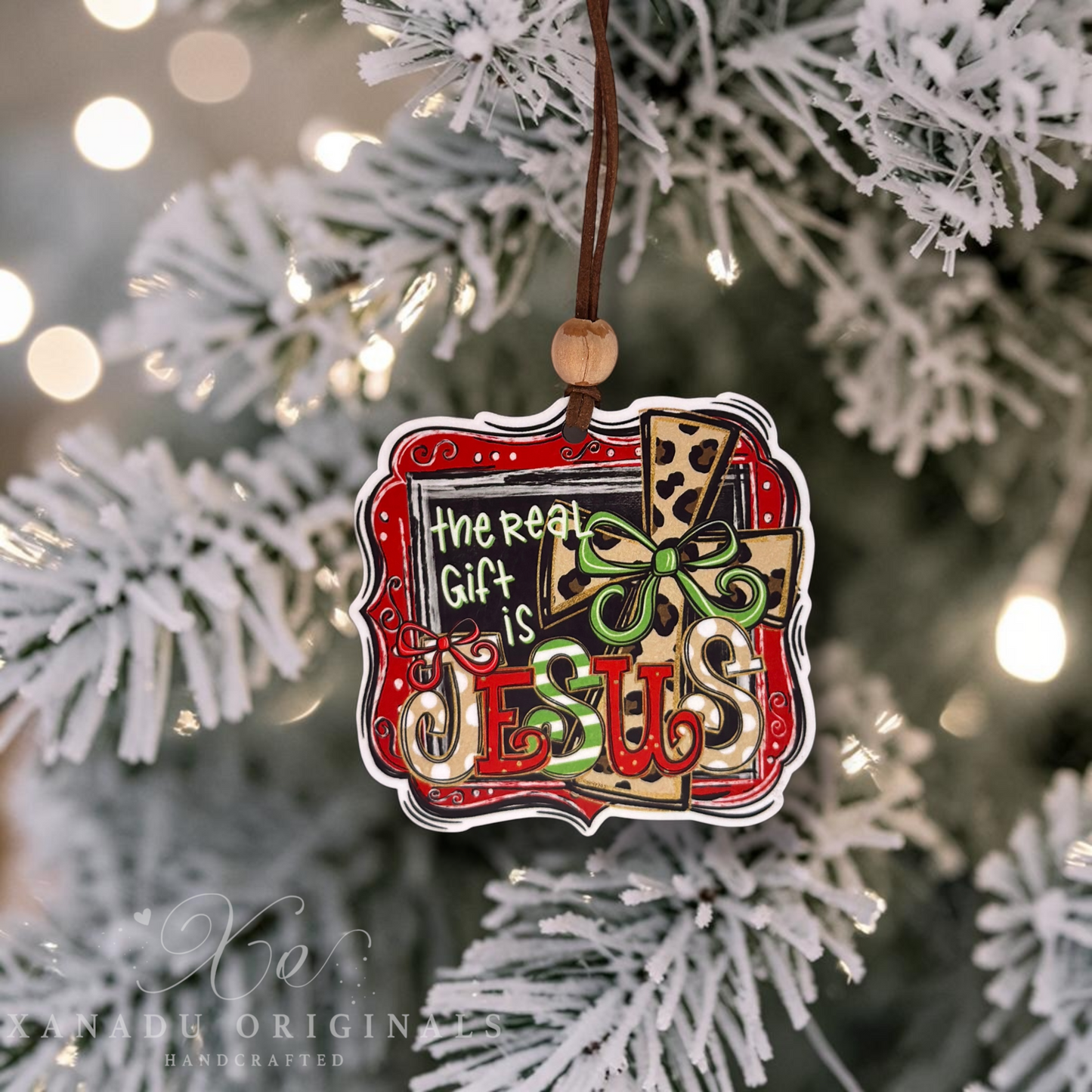 A decorative Christmas ornament hanging from a brown cord on a frosted white Christmas tree. The ornament features the phrase “The Real Gift Is JESUS” in stylized lettering over a gift box design with red, green, beige, and black details.