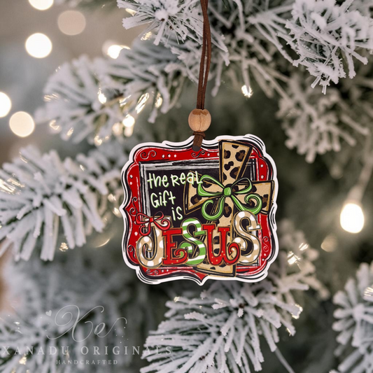 A decorative Christmas ornament hanging from a brown cord on a frosted white Christmas tree. The ornament features the phrase “The Real Gift Is JESUS” in stylized lettering over a gift box design with red, green, beige, and black details.