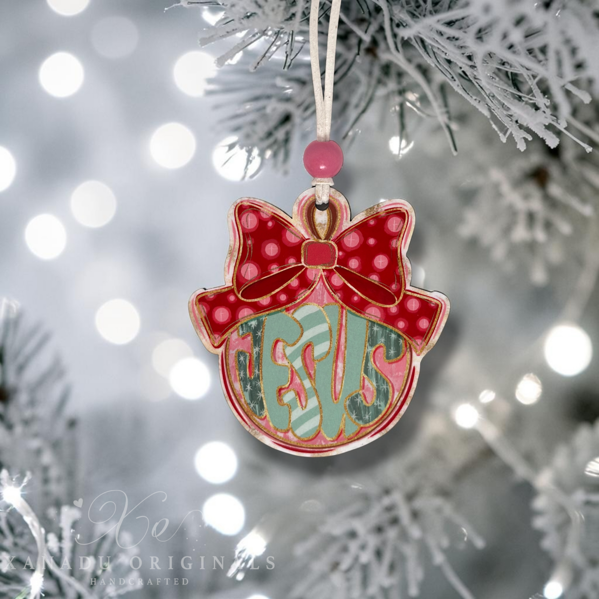 A personalized Christmas ornament shaped like a red bow with the word JESUS, hanging from a white string on a frosted Christmas tree branch with soft bokeh lights in the background.