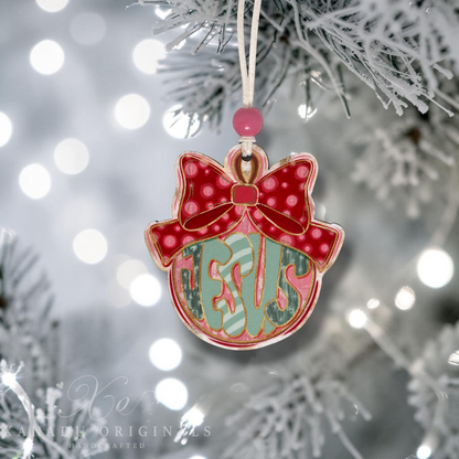 A personalized Christmas ornament shaped like a red bow with the word JESUS, hanging from a white string on a frosted Christmas tree branch with soft bokeh lights in the background.