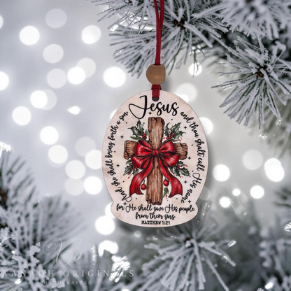 A wooden Christmas ornament with a red hanging ribbon and a wooden bead, featuring a painted wooden cross with a red bow, holly, and scripture text. The ornament is hanging from a frosted Christmas tree branch with soft white bokeh lights in the background.