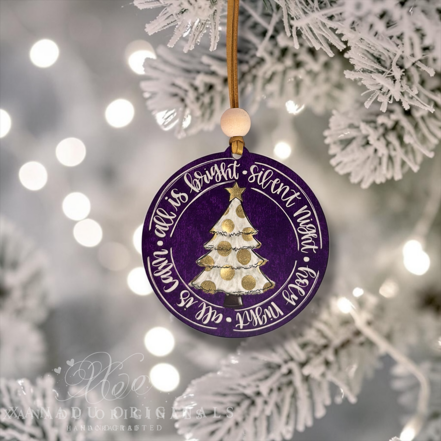A round purple Christmas ornament with a gold hanging string and wooden bead, featuring a white and gold decorated tree and the phrase 'all is quiet - silent night - feast night - all is known' in white script. The ornament is shown hanging among white frosted tree branches with soft bokeh lights in the background.