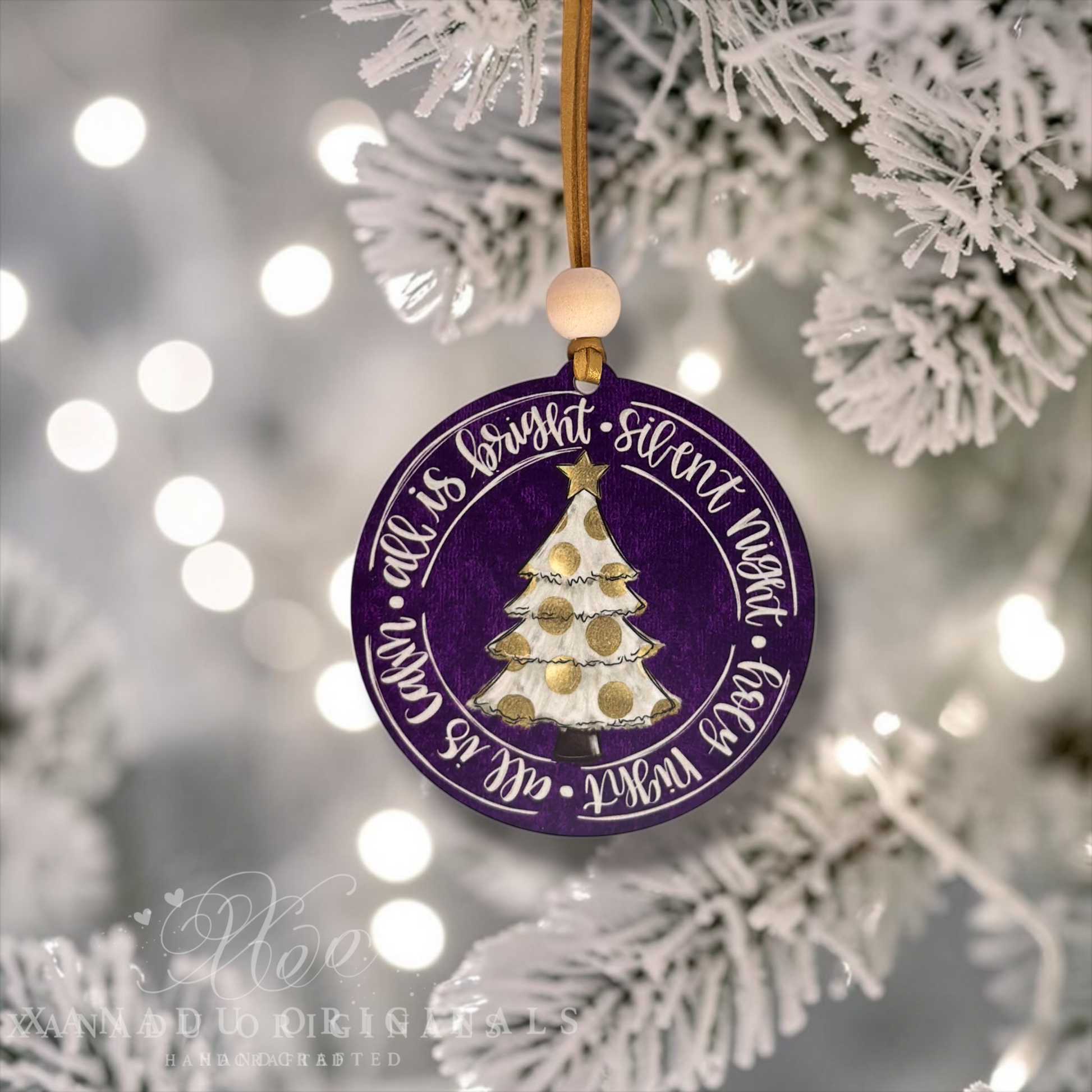 A round purple Christmas ornament with a gold hanging string and wooden bead, featuring a white and gold decorated tree and the phrase 'all is quiet - silent night - feast night - all is known' in white script. The ornament is shown hanging among white frosted tree branches with soft bokeh lights in the background.