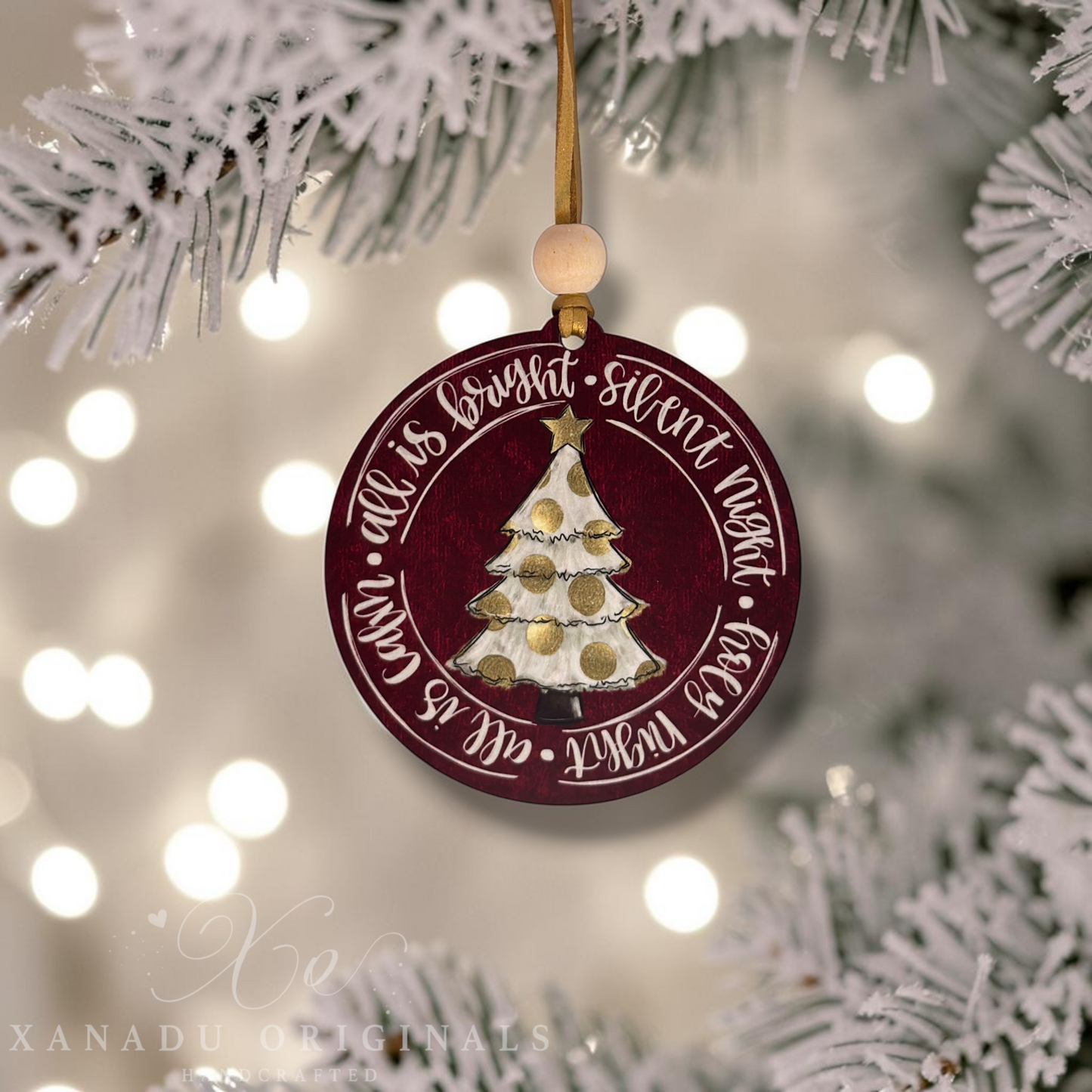 A round Christmas ornament hanging from a gold string, featuring a dark red background with a gold and white decorated Christmas tree in the center and white script text around the edge. The ornament is styled against a blurred festive background with white-frosted tree branches and warm bokeh lights.