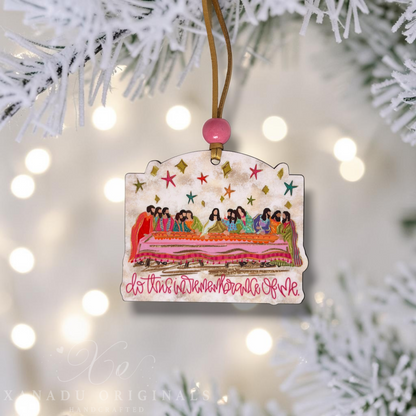 A handcrafted Christmas ornament featuring a colorful illustration of the Last Supper with the text 'do this in remembrance of me' in pink cursive. The ornament is decorated with a gold hanging string, a pink bead, and is shown hanging from white pine tree branches with warm bokeh lights in the background.
