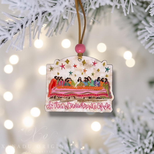 A handcrafted Christmas ornament featuring a colorful illustration of the Last Supper with the text 'do this in remembrance of me' in pink cursive. The ornament is decorated with a gold hanging string, a pink bead, and is shown hanging from white pine tree branches with warm bokeh lights in the background.