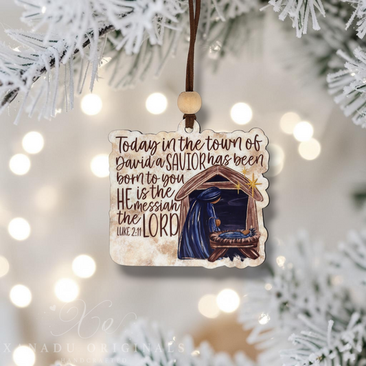 A square Christmas ornament hanging from a brown string with a wooden bead, featuring a nativity scene and the text 'Today in the town of David a SAVIOR has been born to you.