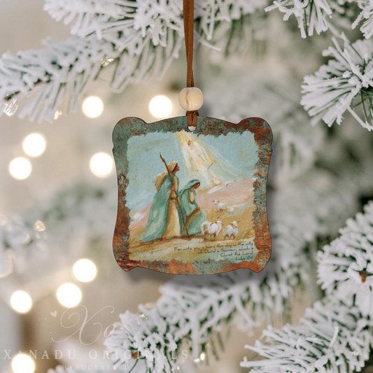 A rustic wood Christmas ornament featuring a painted scene of an Angel with the Two Wise Men and sheep, hanging from a brown ribbon with a wooden bead in front of a blurred flocked Christmas tree.