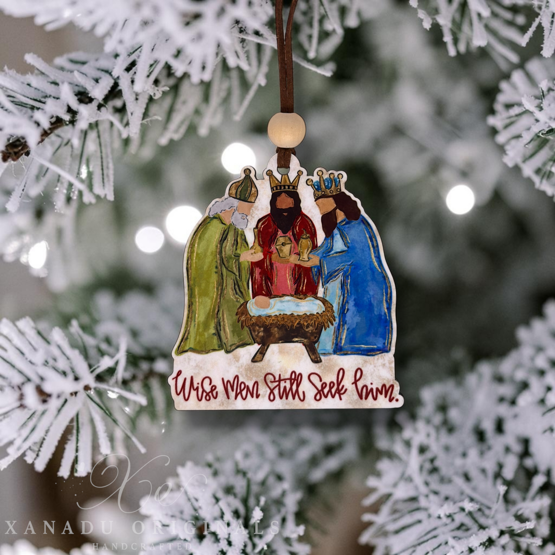 A handcrafted wooden Christmas ornament featuring a painted nativity scene with three wise men, a baby in a manger, and the inscription 'Wise Men Still Seek Love,' hanging from a decorated Christmas tree branch.