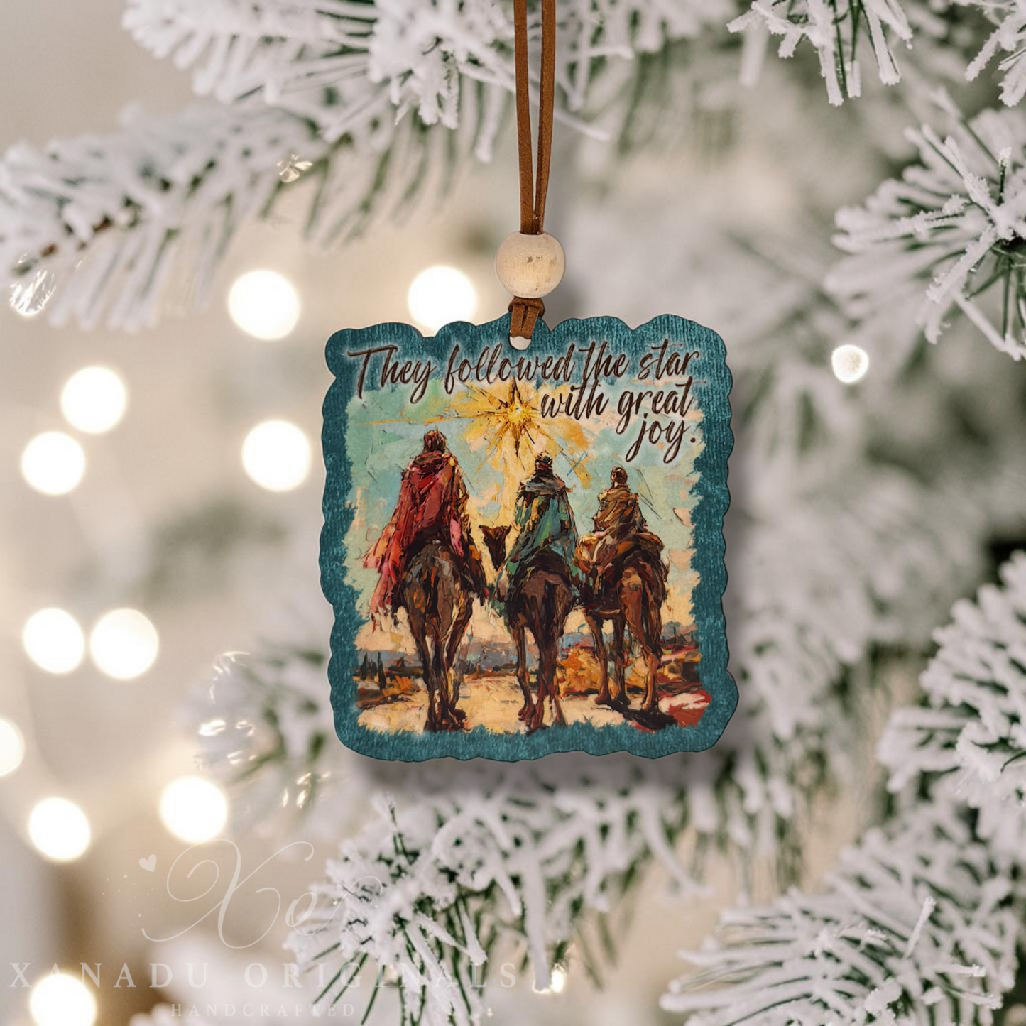 A square Christmas ornament with a rustic teal border hangs from a brown string on a white-frosted Christmas tree. The ornament features an illustration of three camel riders with a glowing star and the text, 'They followed the star with great joy.