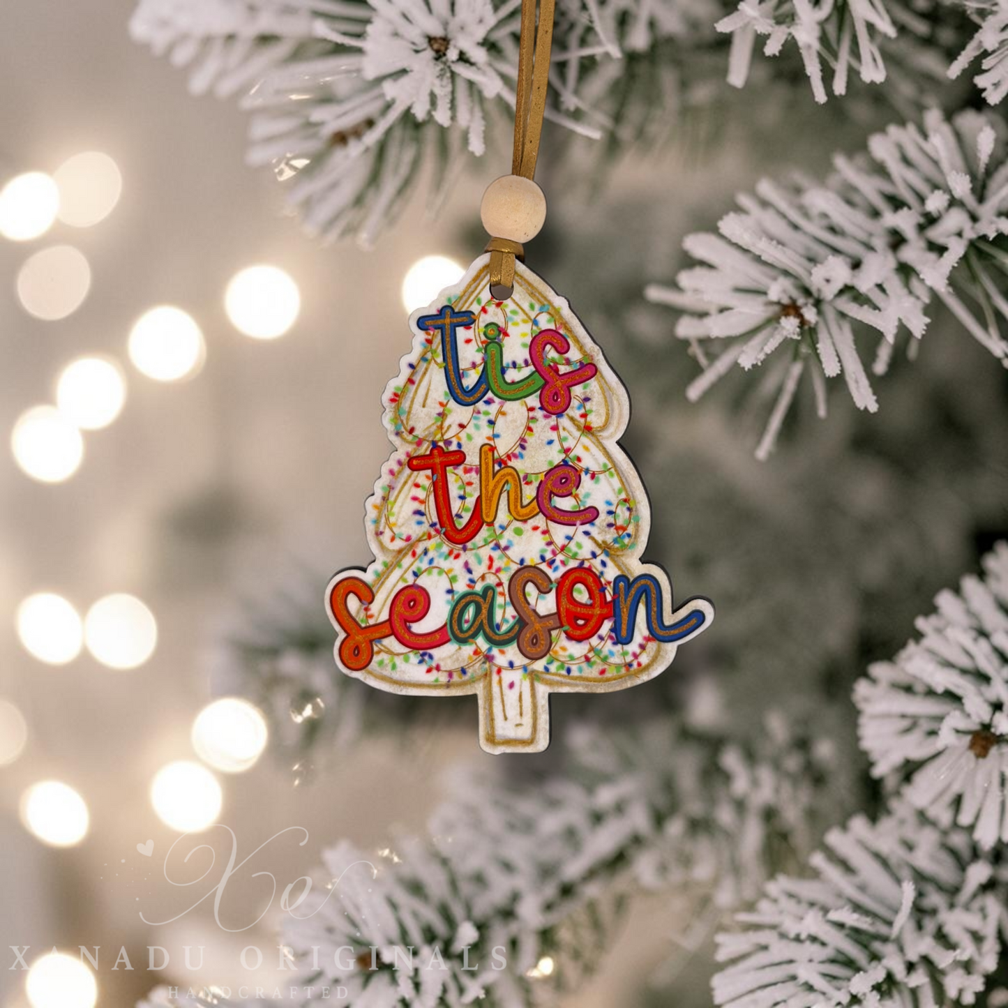 A Christmas tree-shaped ornament hanging from a flocked pine tree branch, with the phrase “tis the season” in colorful script and sprinkle-like detailing. Warm blurred string lights appear in the background.