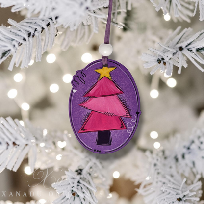 A purple oval Christmas ornament featuring a pink hand-drawn Christmas tree with a yellow star, hanging from a white bead on a string against a blurred white frosted Christmas tree background with warm bokeh lights.