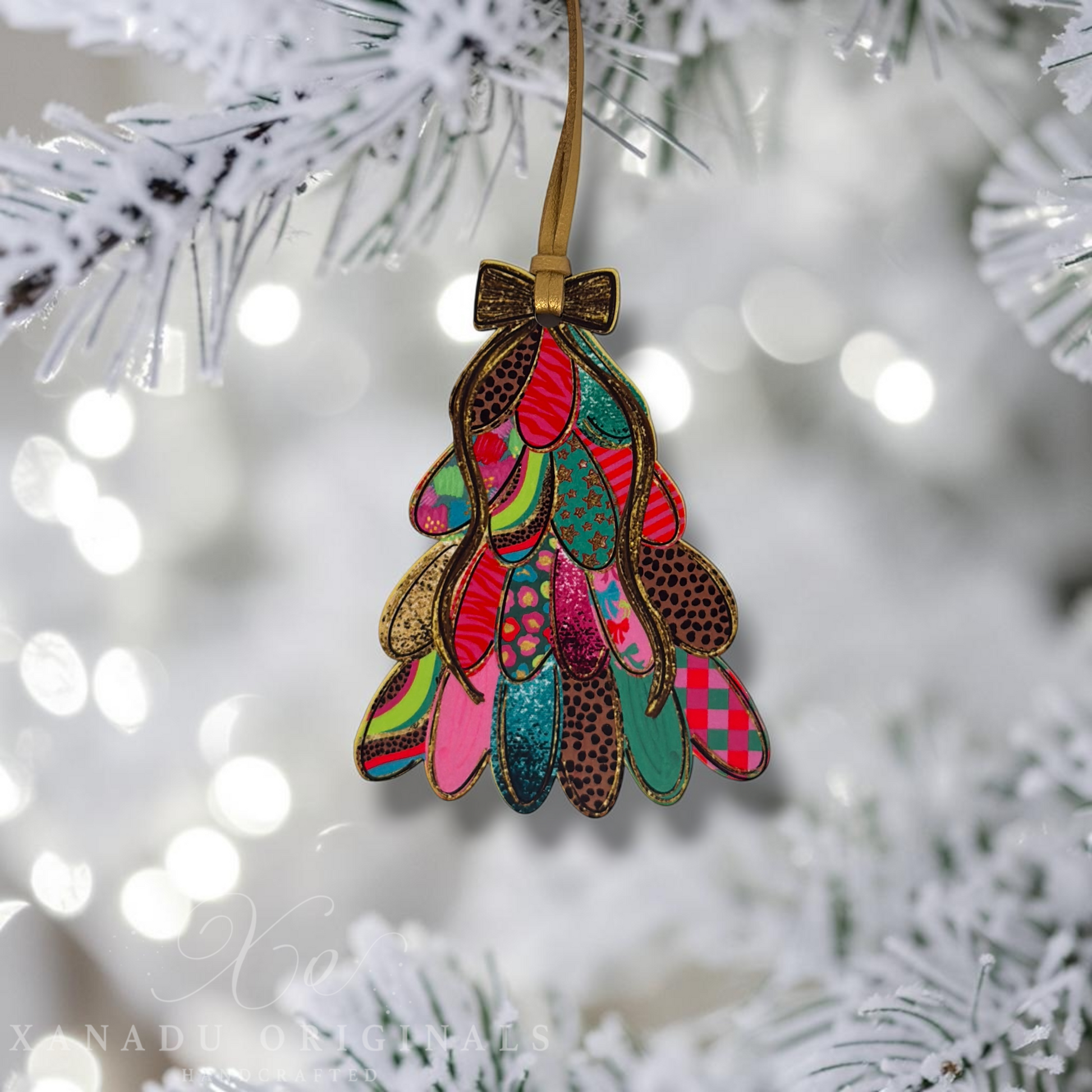 A colorful Christmas tree ornament made of layered patterned paper pieces hangs from a gold ribbon on a white frosted Christmas tree branch with soft bokeh lights in the background.