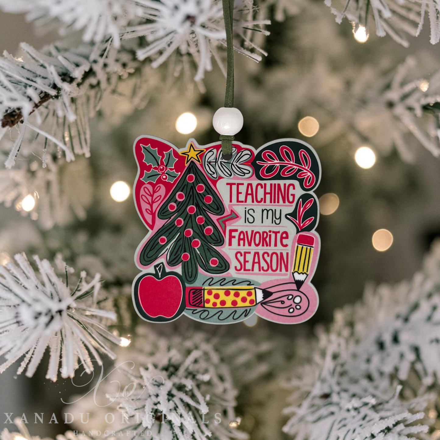 A decorative Christmas ornament shaped like a square sign hangs from a tree branch. The ornament features colorful illustrations of a Christmas tree, an apple, a pencil, a ruler, and the phrase “TEACHING IS MY FAVORITE SEASON” in pink, red, green, black, and white.