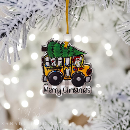A Christmas ornament shaped like a yellow school bus carrying a green Christmas tree, hanging from a gold string on a flocked Christmas tree branch. The ornament has 'Merry Christmas' written at the bottom and a Santa figure in the driver's seat.