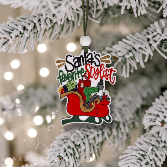 A Christmas ornament shaped like Santa's sleigh with the text 'Santa's favorite stylist' hanging from a white-frosted Christmas tree branch.