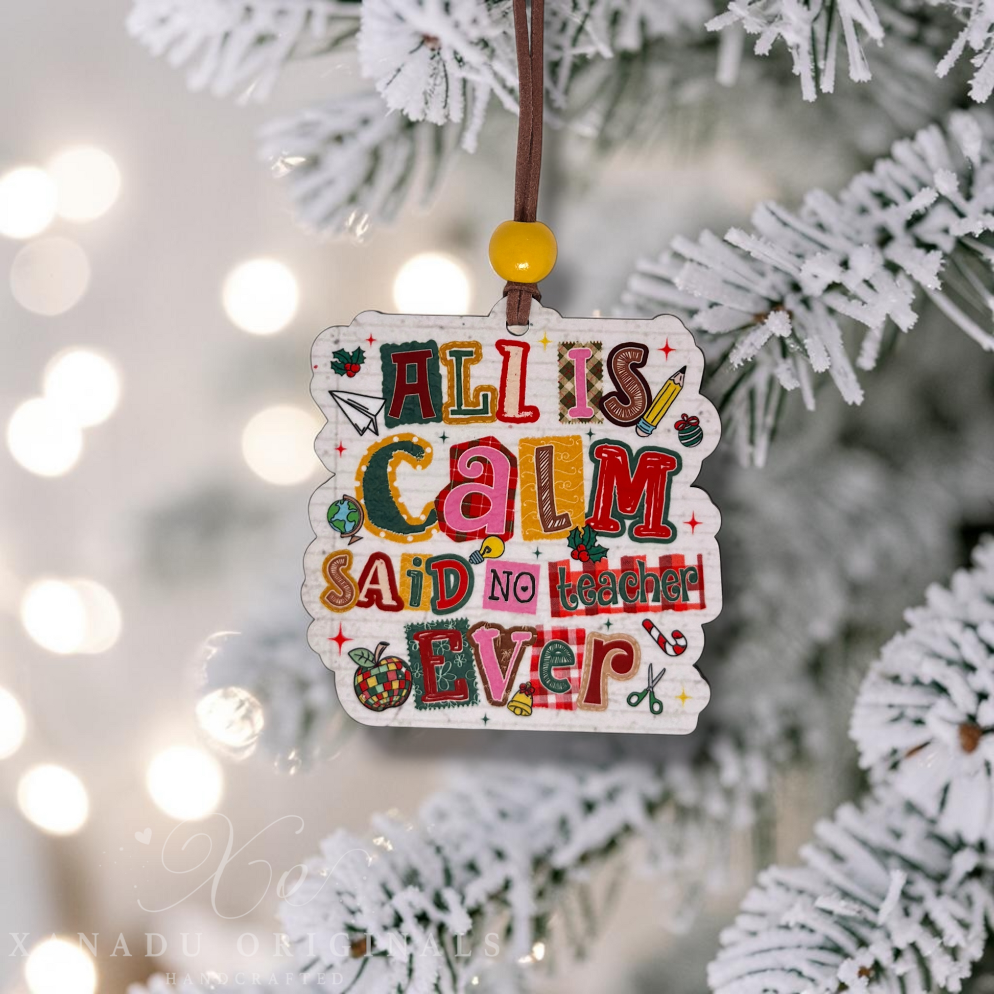 A decorative Christmas ornament shaped like a white sign with colorful hand-lettered text reading 'ALL IS CALM SAID NO TEACHER EVER,' hanging from a brown cord with a yellow bead on a blurred white-frosted Christmas tree branch.