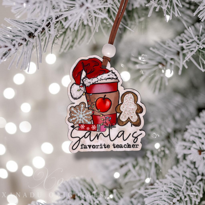 A Christmas ornament hanging from a flocked pine tree branch. The ornament features a coffee cup wearing a Santa hat, gingerbread cookies, a red apple, and the text 'Santa's favorite teacher' in black script.