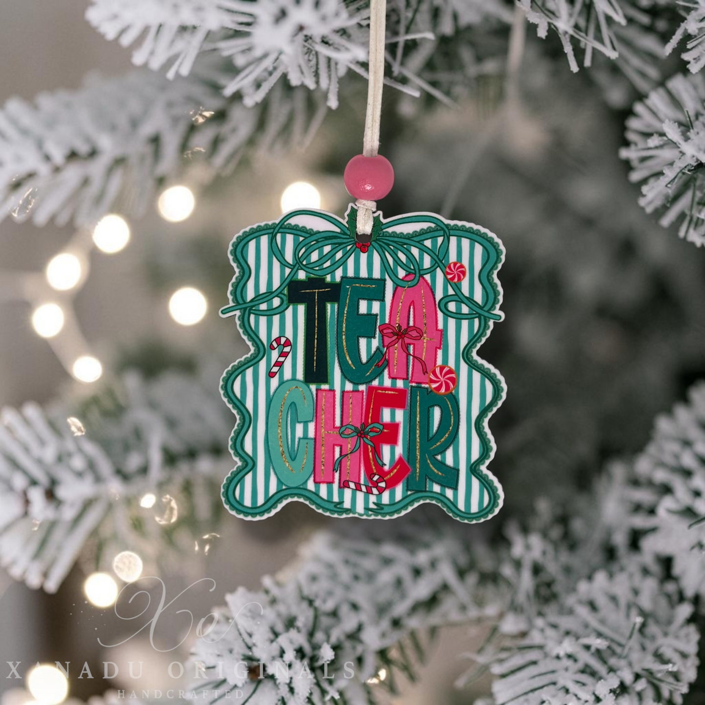 A decorative Christmas ornament shaped like a gift box with the word "TEACHER" in colorful letters, hanging from a white string on a frosted Christmas tree with warm string lights in the background.