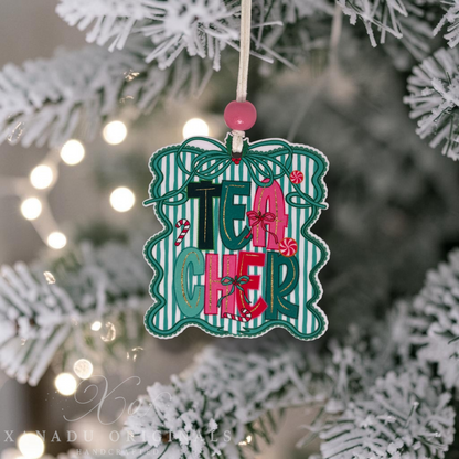 A decorative Christmas ornament shaped like a gift box with the word "TEACHER" in colorful letters, hanging from a white string on a frosted Christmas tree with warm string lights in the background.