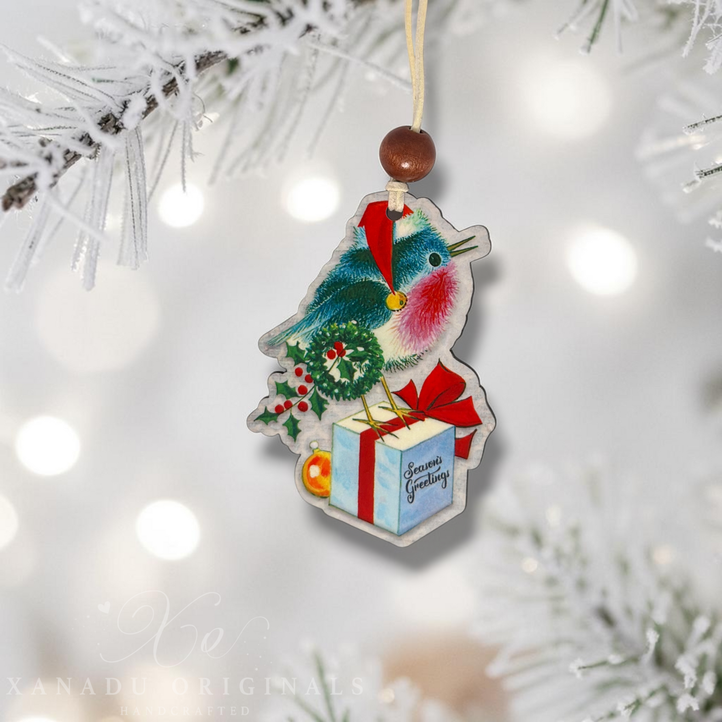A Christmas bird ornament hanging from a white-frosted tree branch. The ornament features a bluebird wearing a Santa hat and scarf, perched on a small wreath next to a wrapped gift box with a red bow and a gold ornament.