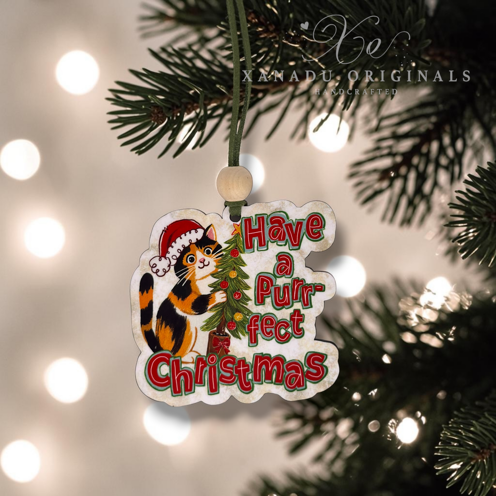 A Christmas ornament shaped like a cat wearing a Santa hat, hanging from a green tree branch with warm blurred lights in the background. The ornament reads 'Have a Purrfect Christmas' in red and green lettering.