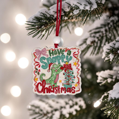 A square Christmas ornament with a scalloped edge hangs from a red ribbon on a snowy tree branch. The ornament features a green alligator wearing a Santa hat, a decorated Christmas tree, and the text 'Have a Snappy Christmas' in colorful lettering.