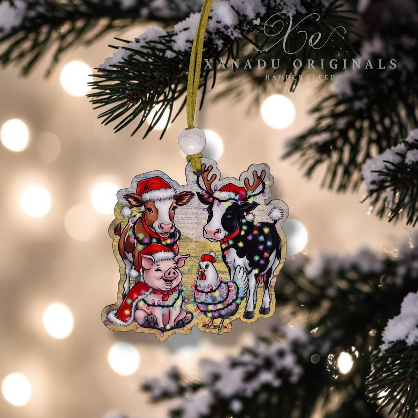 A Christmas ornament featuring illustrated farm animals dressed in holiday attire, hanging from a gold ribbon on a snow-dusted evergreen tree with warm blurred lights in the background. The ornament has a decorative border and a visible 'XNADU ORIGINALS HANDCRAFTED' watermark/logo in the upper right.