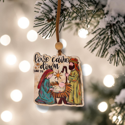 A hand-painted wooden Christmas ornament featuring a nativity scene with Mary, Joseph, baby Jesus, and a star, hanging from a wooden string with a bead. The ornament is displayed against a blurred background of a Christmas tree and warm bokeh lights.