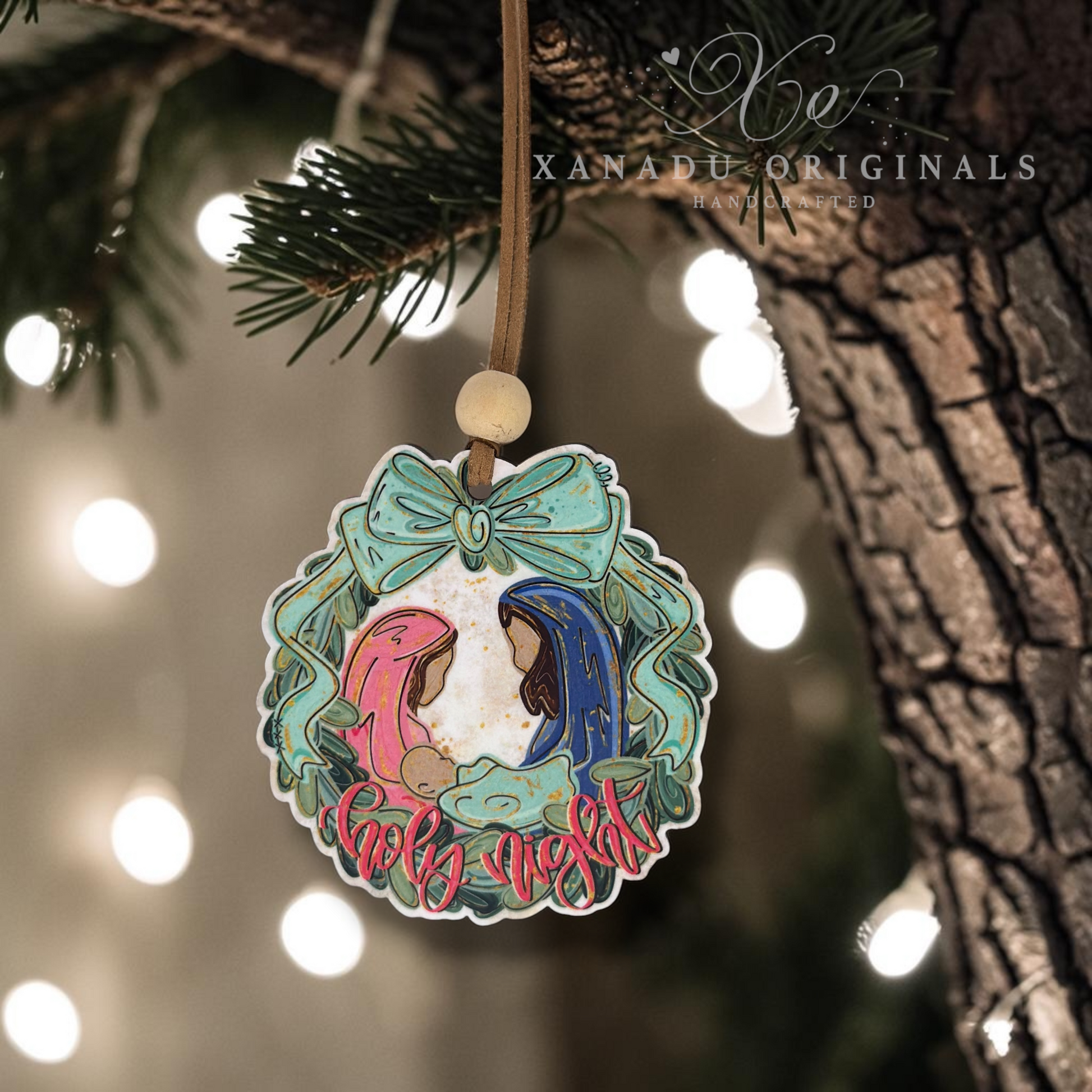 A round Christmas ornament hanging from a tree branch with warm string lights in the background. The ornament features an illustrated nativity scene with Mary, Joseph, baby Jesus, and a pink donkey, surrounded by a mint green wreath and bow, with the words 'Holy Night' at the bottom.