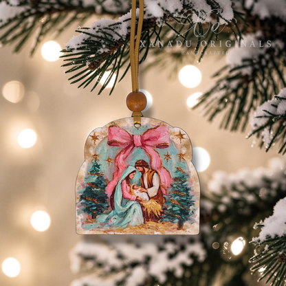 A hand-painted Christmas ornament depicting the Holy Family under a large pink bow, hanging from a pine tree branch with snowy lights in the background.