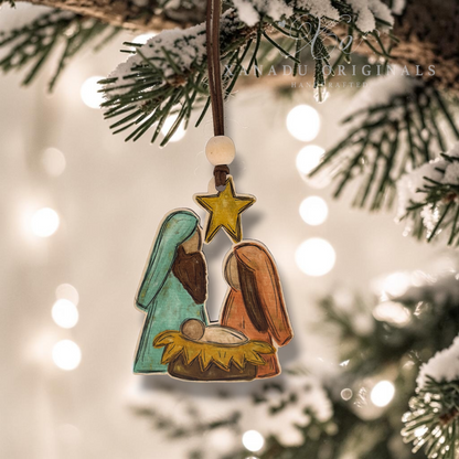 Hand-painted wooden Nativity ornament hanging from a snowy Christmas tree branch with warm bokeh lights in the background. The ornament features Mary in teal, Joseph in brown, baby Jesus in a yellow manger, and a gold star above.