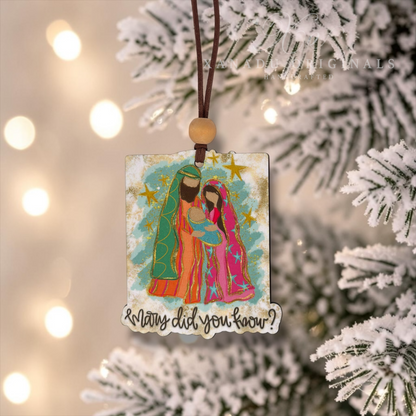 A rectangular Christmas ornament with a colorful illustration of Mary and Joseph under gold stars, hanging from a brown cord with a wooden bead. The ornament is displayed on a snowy Christmas tree with warm bokeh lights in the background.
