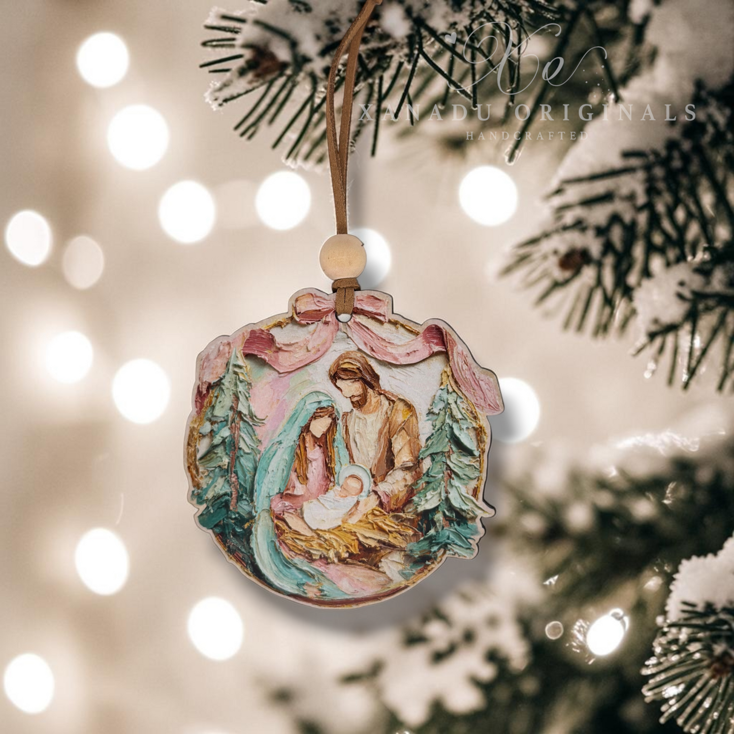 A round hand-painted Christmas ornament depicting the Holy Family under a pink bow, hanging from a snowy Christmas tree branch with warm bokeh lights in the background.
