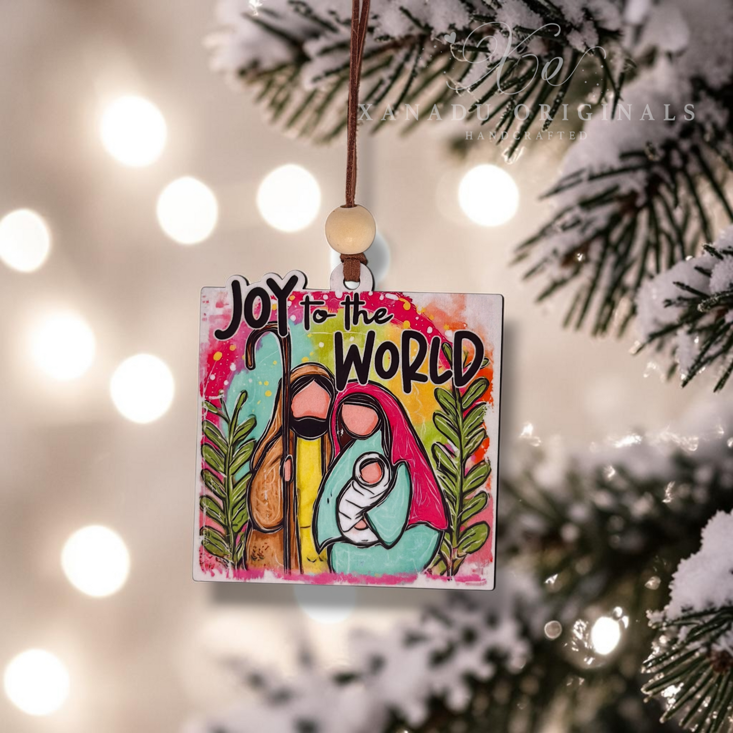 A square wooden Christmas ornament hanging from a brown string with a wooden bead, featuring a colorful painted illustration of Mary, Joseph, and baby Jesus with the text 'JOY to the WORLD'. The ornament is photographed hanging in front of a blurred Christmas tree and warm bokeh lights.