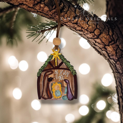 A hand-painted wooden Christmas ornament shaped like a stable, hanging from a tree branch. The ornament features a brown wood-grain background with a painted nativity scene including a yellow star, green wreath, and figures in blue and purple.