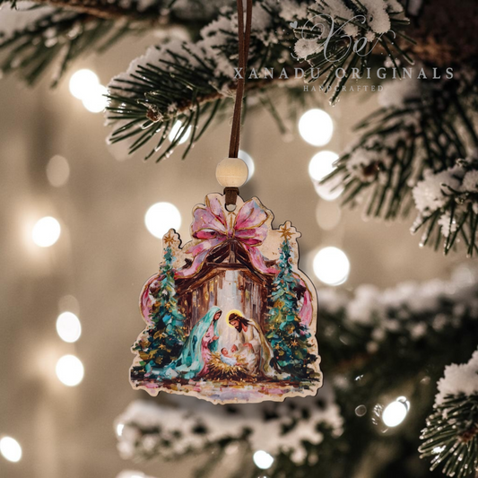 A Christmas nativity ornament featuring Mary, Joseph, and baby Jesus under a pink bow, hanging from a pine tree branch with warm bokeh lights in the background. The brand name 'Xanadu Originals' is watermarked at the top right.